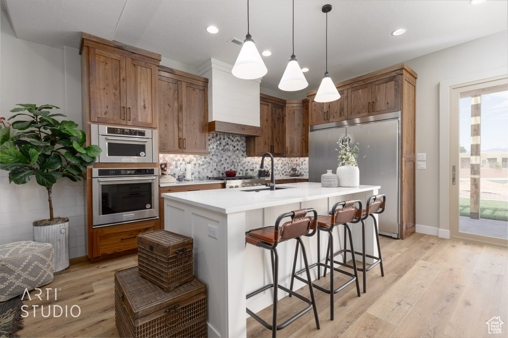 Kitchen with light wood-type flooring, custom range hood, appliances with stainless steel finishes, an island with sink, and decorative light fixtures