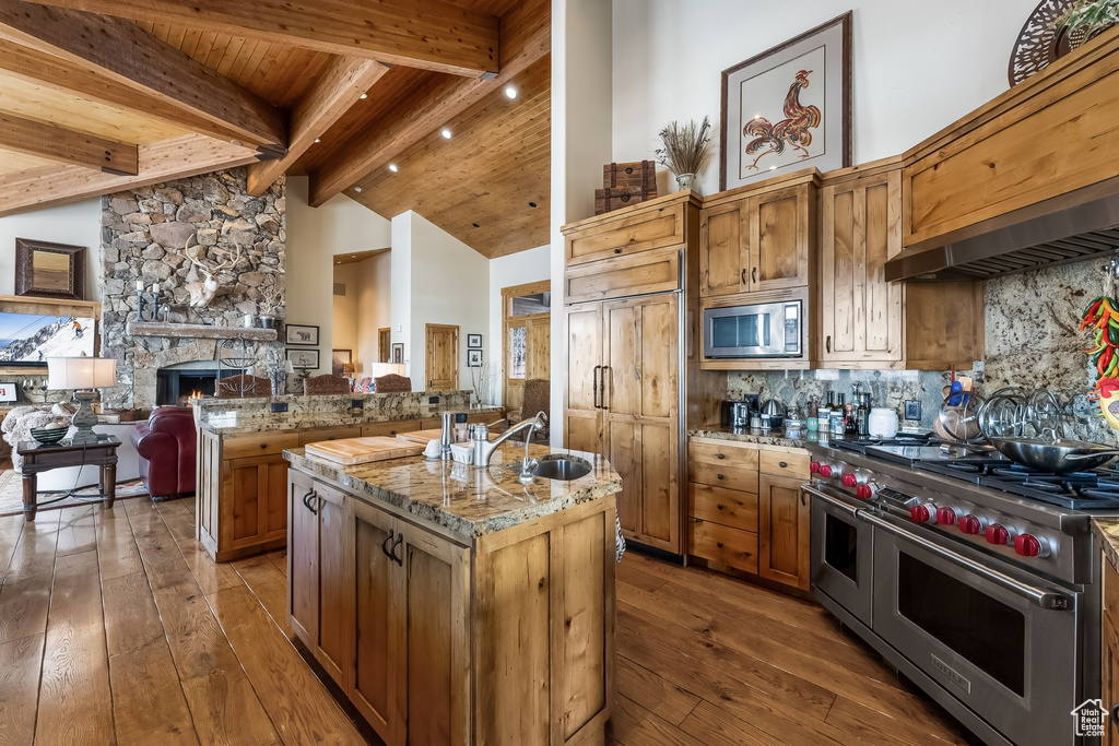 Kitchen with appliances with stainless steel finishes, a kitchen island with sink, a fireplace, beam ceiling, and dark hardwood / wood-style floors