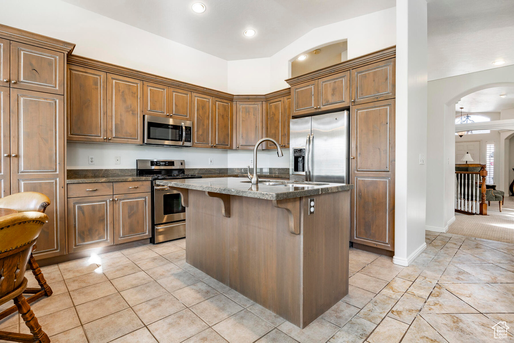 Kitchen featuring a kitchen bar, an island with sink, dark stone countertops, appliances with stainless steel finishes, and light colored carpet