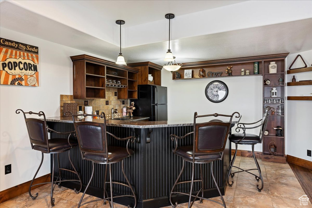 Bar with backsplash, black refrigerator, light tile floors, decorative light fixtures, and dark brown cabinetry
