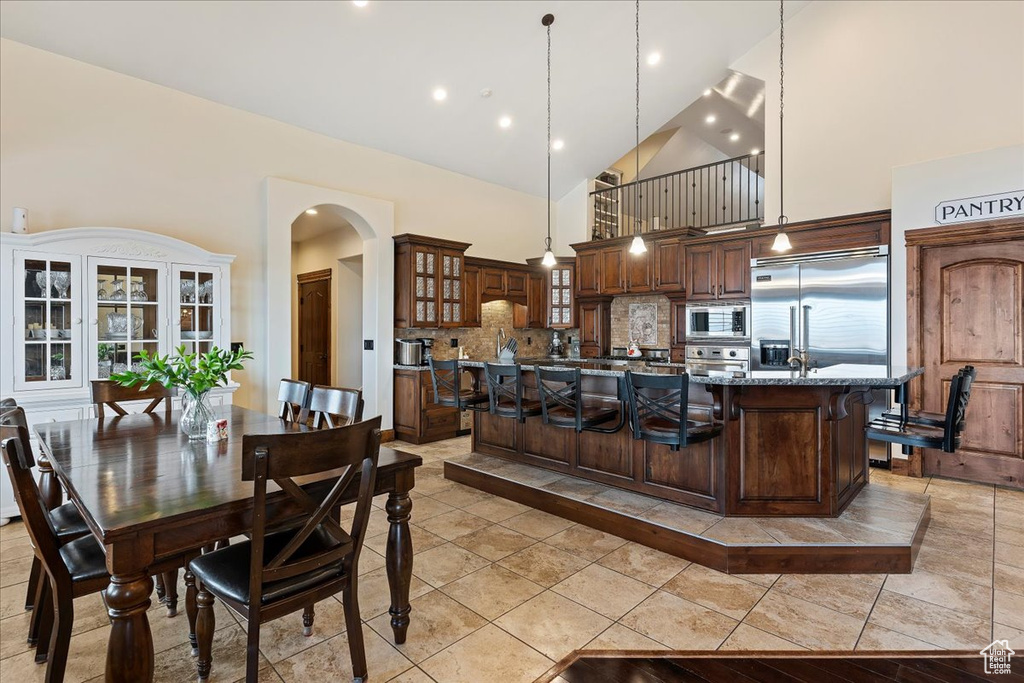 Kitchen with built in appliances, dark stone counters, a breakfast bar, backsplash, and decorative light fixtures