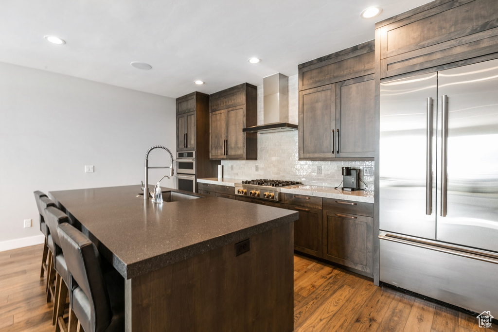 Kitchen with tasteful backsplash, dark hardwood / wood-style floors, an island with sink, appliances with stainless steel finishes, and wall chimney exhaust hood