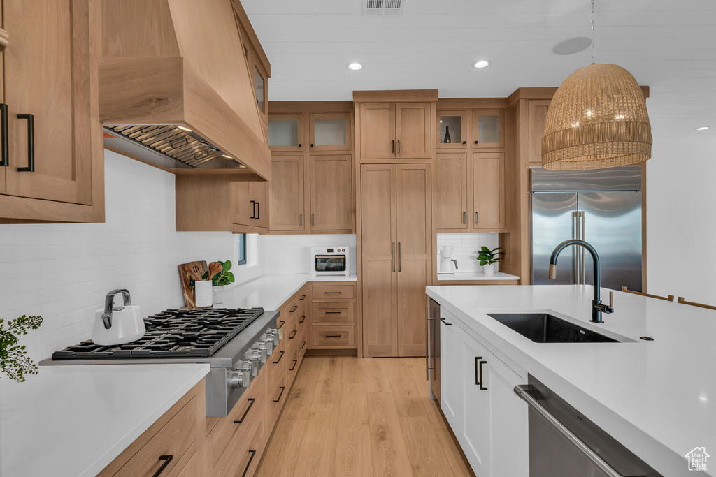Kitchen with light hardwood / wood-style floors, hanging light fixtures, custom exhaust hood, sink, and built in appliances