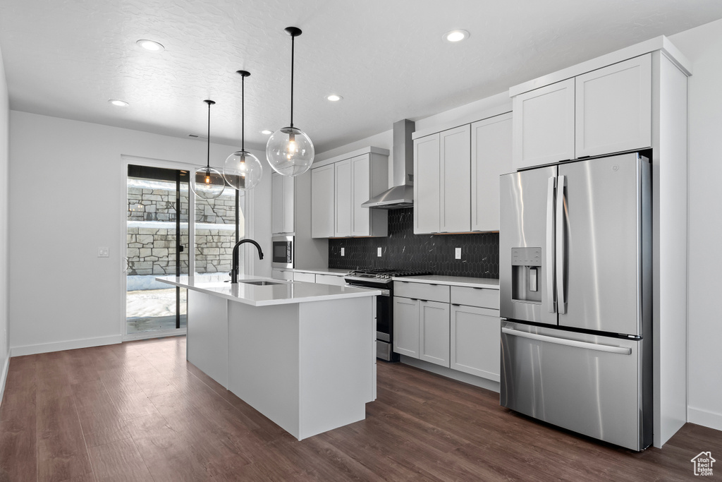 Kitchen featuring pendant lighting, dark wood-type flooring, appliances with stainless steel finishes, wall chimney range hood, and a kitchen island with sink