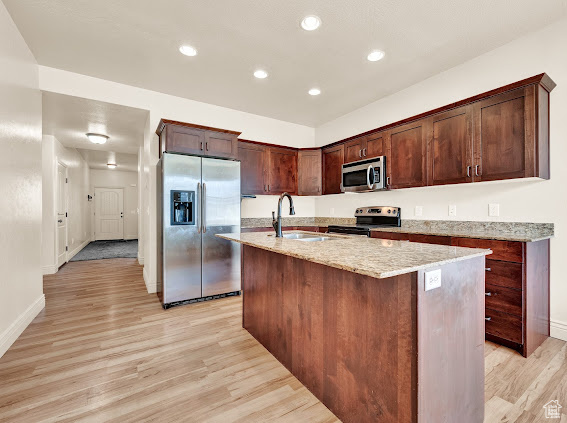 Kitchen with light stone countertops, light hardwood / wood-style floors, appliances with stainless steel finishes, and an island with sink