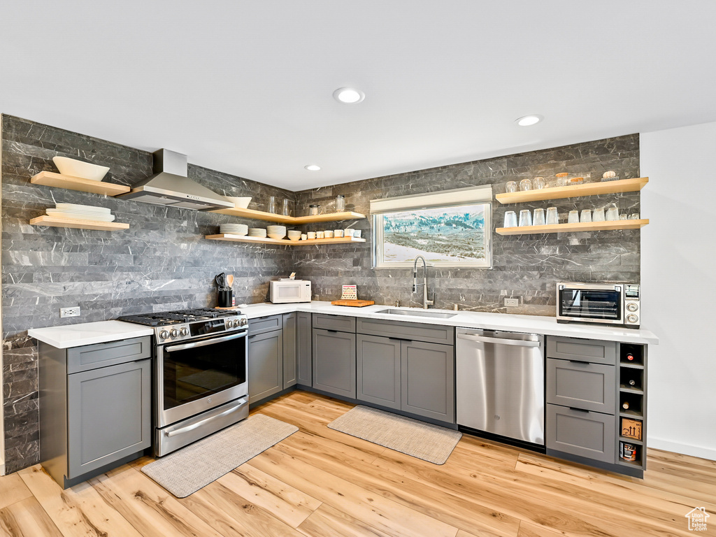 Kitchen with wall chimney exhaust hood, light wood-type flooring, gray cabinets, stainless steel appliances, and sink