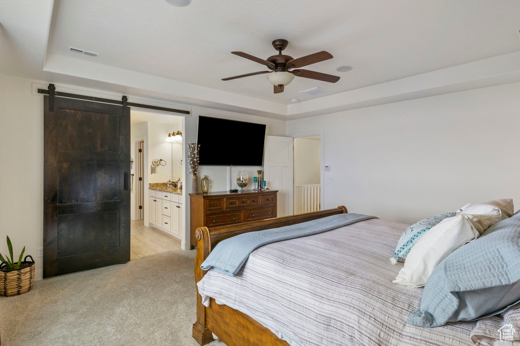 Bedroom with a raised ceiling, a barn door, light colored carpet, connected bathroom, and ceiling fan