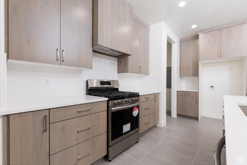 Kitchen with stainless steel gas range oven, light tile patterned floors, decorative backsplash, light brown cabinets, and wall chimney range hood