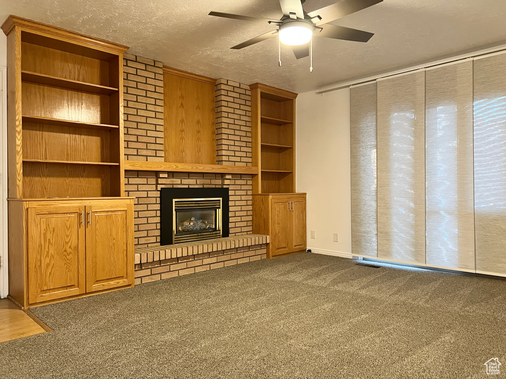 Unfurnished living room with a textured ceiling, a fireplace, brick wall, ceiling fan, and dark colored carpet