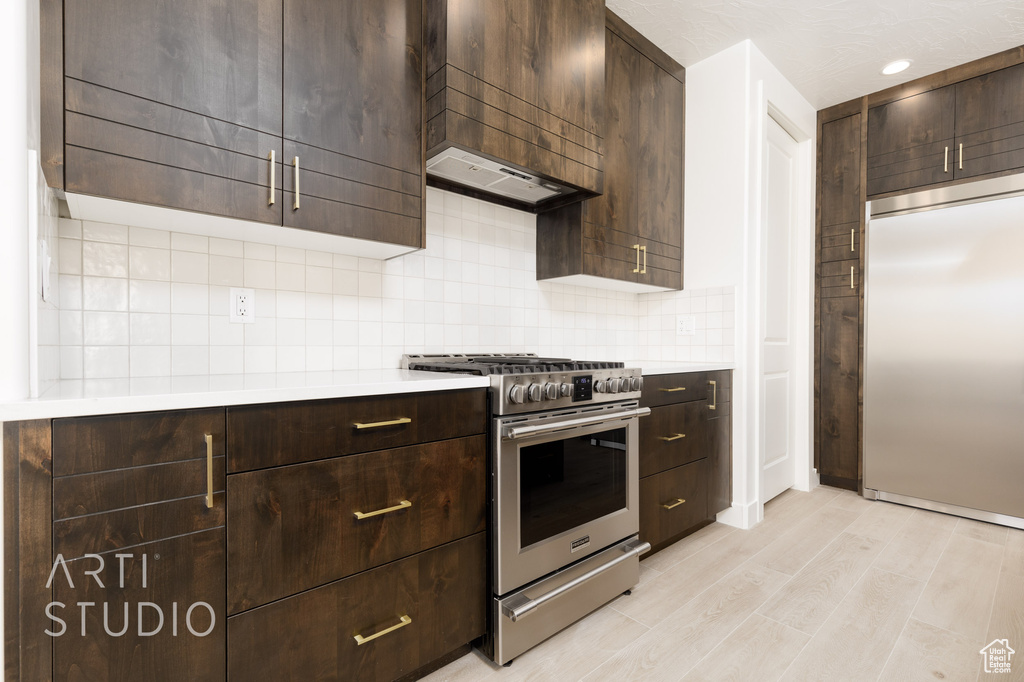 Kitchen with tasteful backsplash, high end appliances, ventilation hood, light hardwood / wood-style floors, and dark brown cabinets