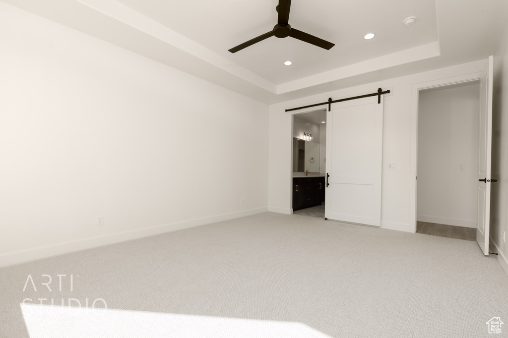 Unfurnished bedroom featuring ceiling fan, a raised ceiling, light carpet, connected bathroom, and a barn door