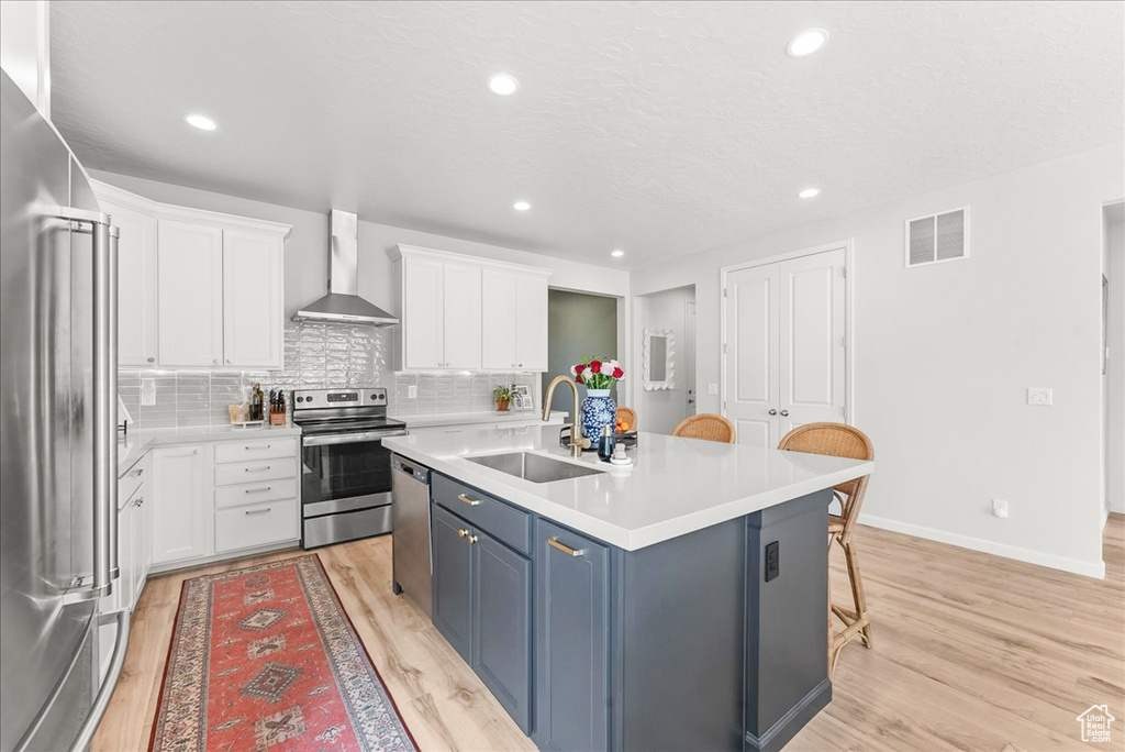 Kitchen featuring light hardwood / wood-style floors, a center island with sink, stainless steel appliances, and wall chimney exhaust hood