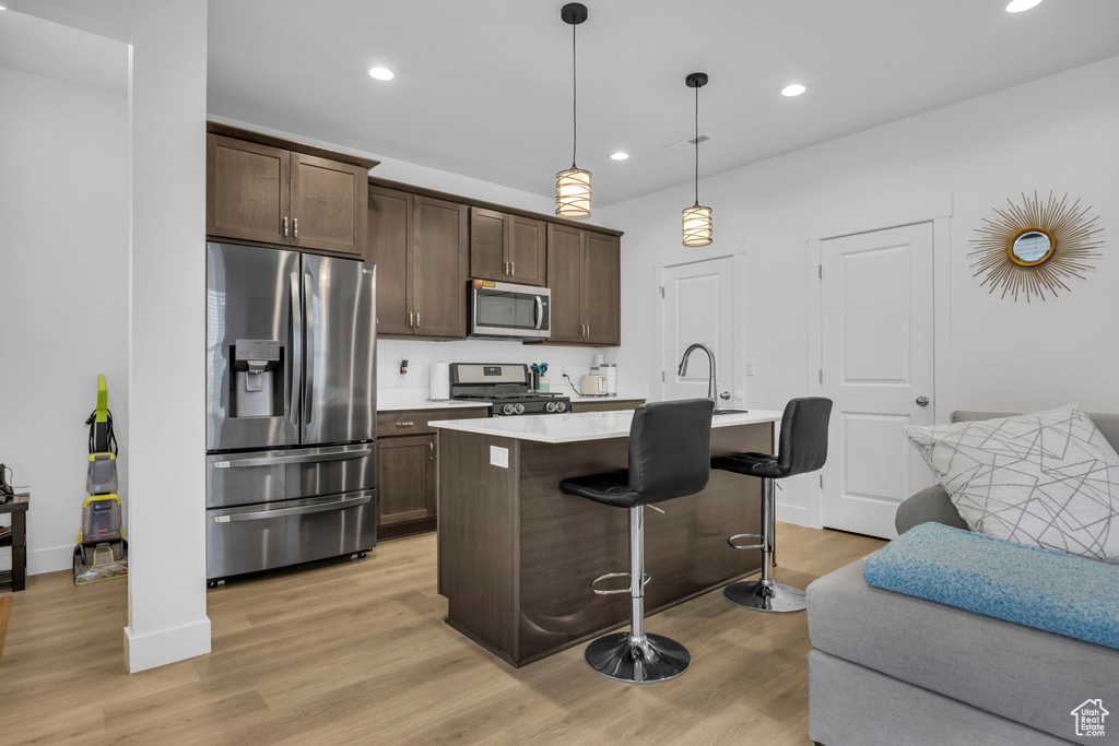 Kitchen with pendant lighting, dark brown cabinets, stainless steel appliances, light hardwood / wood-style flooring, and a kitchen island with sink