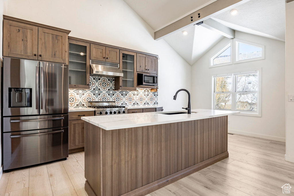 Kitchen featuring appliances with stainless steel finishes, light hardwood / wood-style floors, an island with sink, beam ceiling, and tasteful backsplash