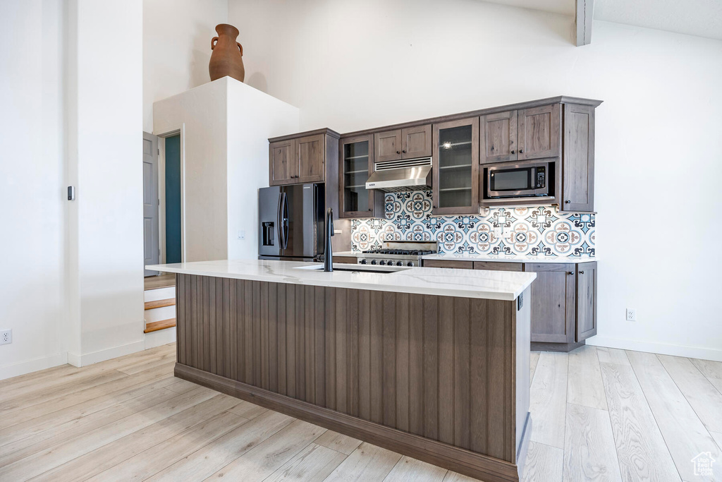 Kitchen with backsplash, a kitchen island with sink, appliances with stainless steel finishes, high vaulted ceiling, and light hardwood / wood-style flooring