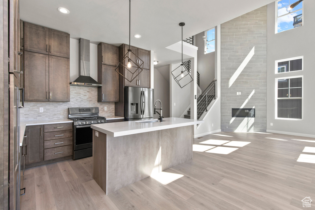 Kitchen featuring light wood-type flooring, a healthy amount of sunlight, a high ceiling, appliances with stainless steel finishes, and wall chimney range hood