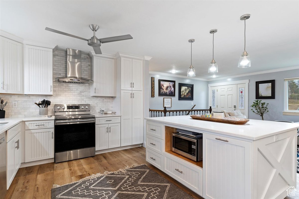 Kitchen featuring light hardwood / wood-style flooring, ceiling fan, decorative light fixtures, appliances with stainless steel finishes, and wall chimney range hood