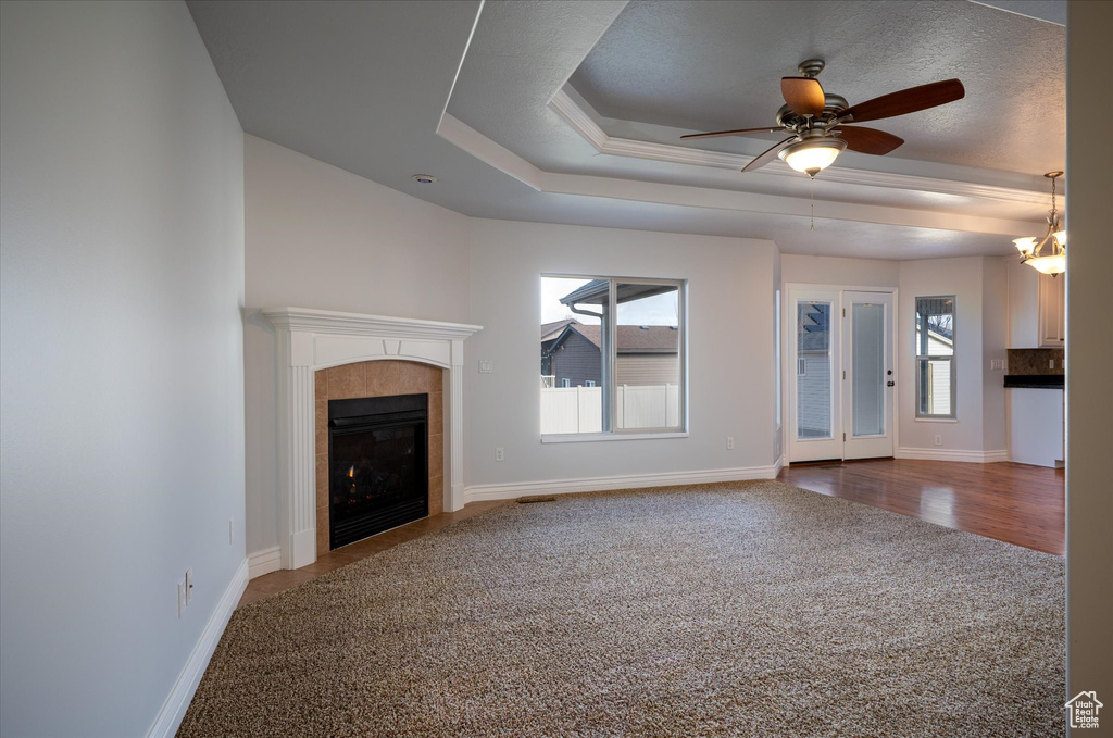 Unfurnished living room featuring a tiled fireplace, dark wood-type flooring, ceiling fan with notable chandelier, and a tray ceiling