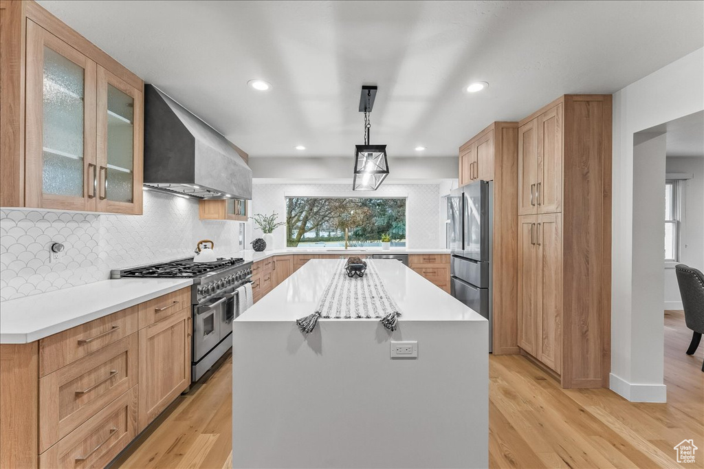 Kitchen with light hardwood / wood-style flooring, a kitchen island, hanging light fixtures, appliances with stainless steel finishes, and wall chimney exhaust hood