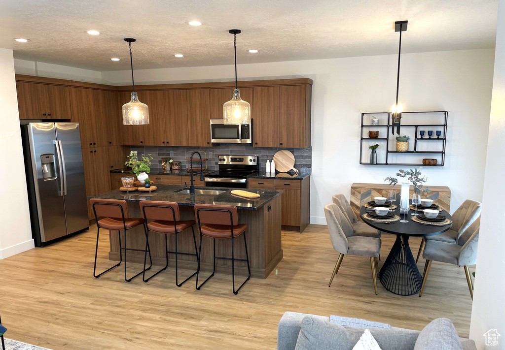 Kitchen featuring light hardwood / wood-style flooring, an island with sink, stainless steel appliances, and decorative light fixtures