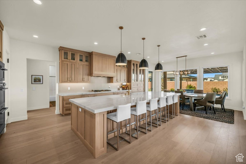 Kitchen with light hardwood / wood-style floors, hanging light fixtures, custom range hood, an island with sink, and a notable chandelier