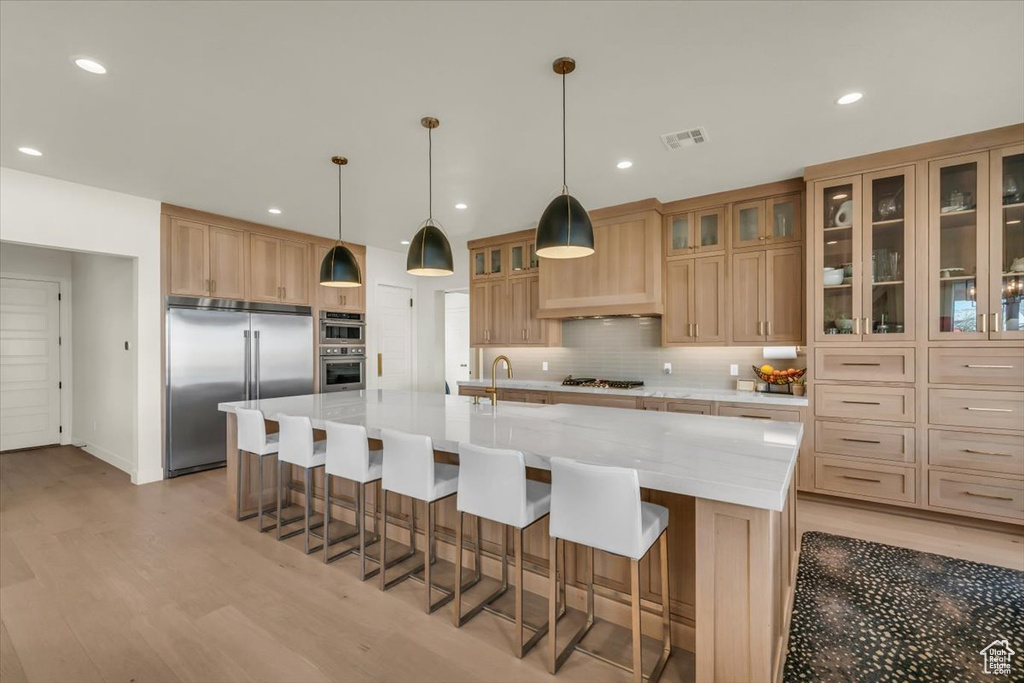 Kitchen with light wood-type flooring, appliances with stainless steel finishes, pendant lighting, and a kitchen island with sink