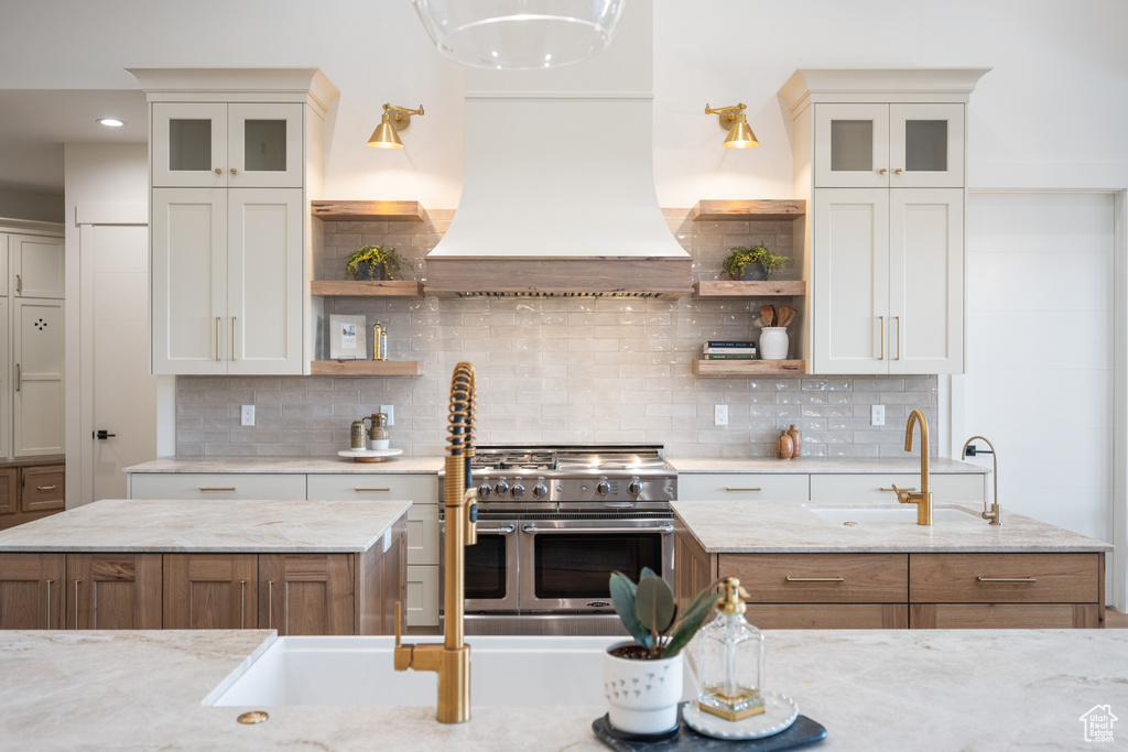 Kitchen featuring custom exhaust hood, light stone countertops, range with two ovens, and backsplash