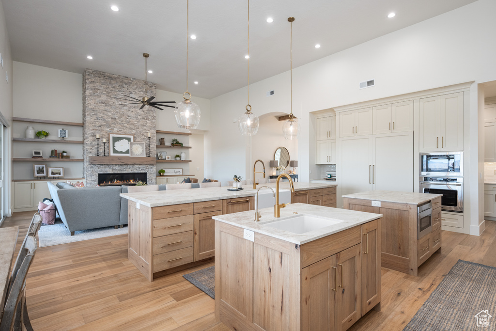 Kitchen with a stone fireplace, a center island with sink, appliances with stainless steel finishes, light hardwood / wood-style flooring, and ceiling fan