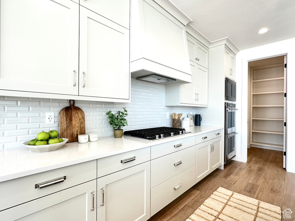 Kitchen with light hardwood / wood-style floors, tasteful backsplash, white cabinetry, custom range hood, and stainless steel appliances