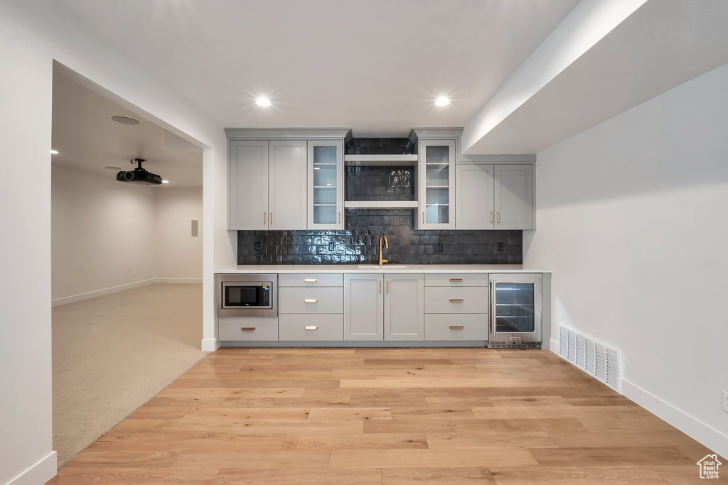 Bar featuring backsplash, stainless steel microwave, beverage cooler, gray cabinetry, and light carpet