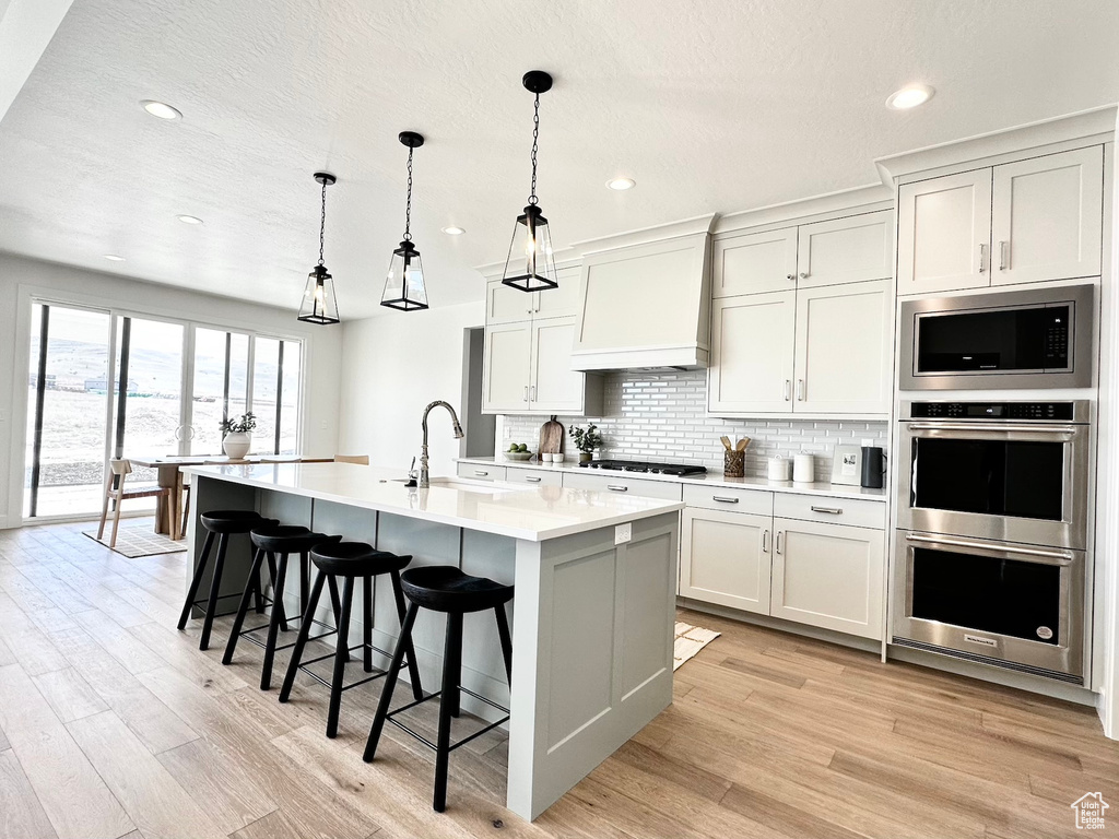 Kitchen with appliances with stainless steel finishes, premium range hood, light hardwood / wood-style flooring, a kitchen island with sink, and hanging light fixtures