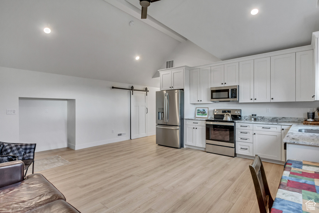 Kitchen with stainless steel appliances, high vaulted ceiling, white cabinetry, a barn door, and light hardwood / wood-style flooring