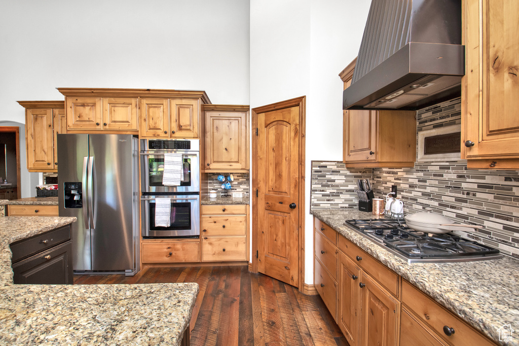 Kitchen featuring wall chimney exhaust hood, dark hardwood / wood-style flooring, light stone countertops, backsplash, and appliances with stainless steel finishes