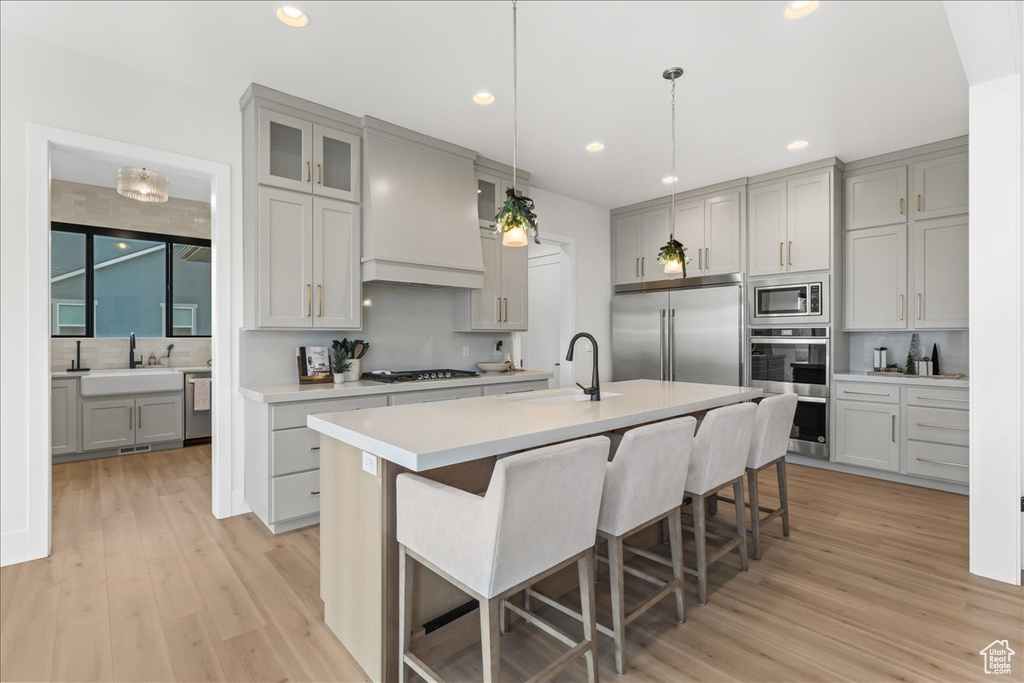 Kitchen featuring built in appliances, light hardwood / wood-style flooring, custom exhaust hood, and a kitchen island with sink