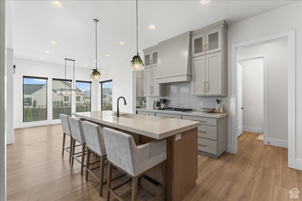 Kitchen with custom exhaust hood, light hardwood / wood-style flooring, gray cabinetry, and a center island with sink