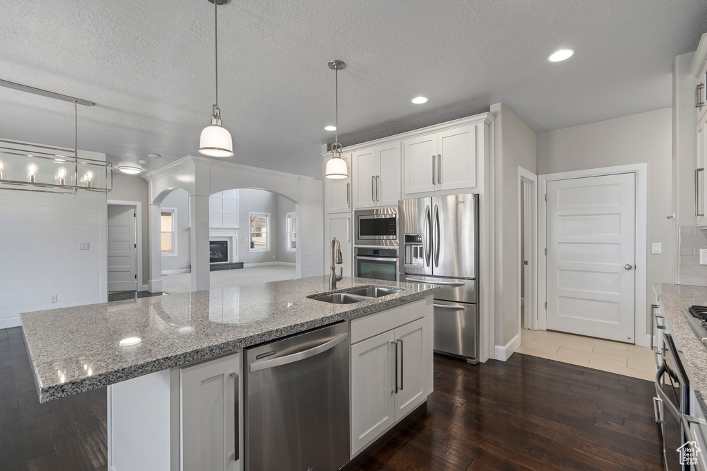 Kitchen featuring dark wood-type flooring, white cabinetry, appliances with stainless steel finishes, an island with sink, and decorative light fixtures