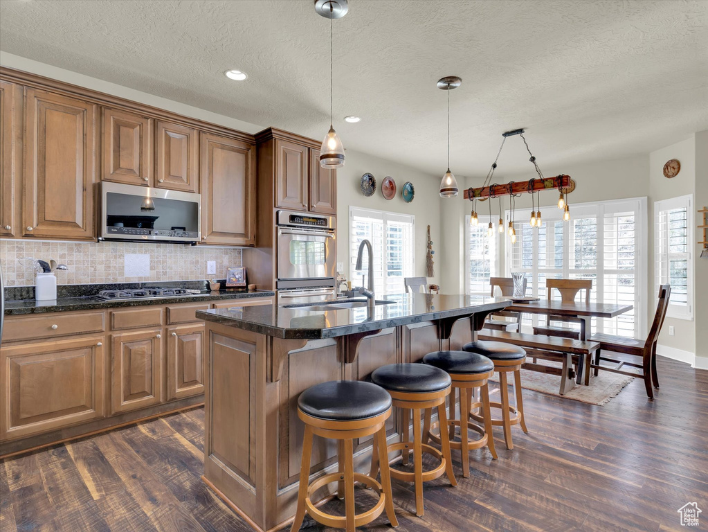 Kitchen with dark hardwood / wood-style floors, appliances with stainless steel finishes, a center island with sink, and decorative light fixtures