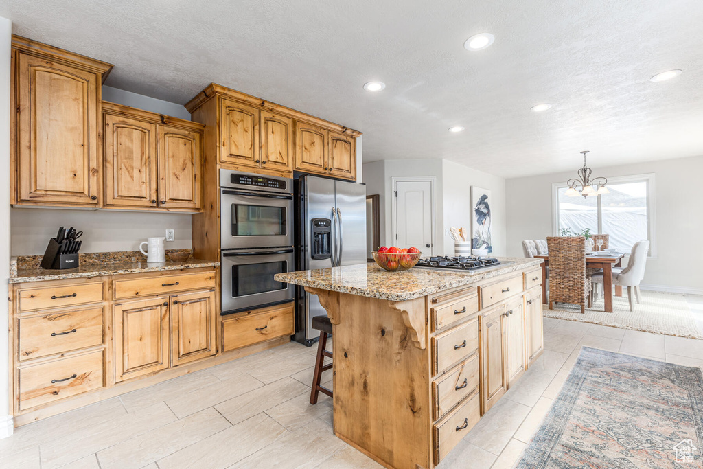 Kitchen with light stone countertops, appliances with stainless steel finishes, a notable chandelier, a center island, and decorative light fixtures