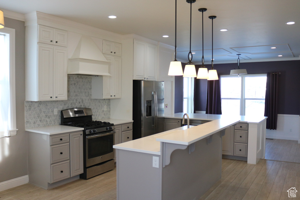 Kitchen with custom range hood, a center island with sink, appliances with stainless steel finishes, light hardwood / wood-style flooring, and decorative light fixtures