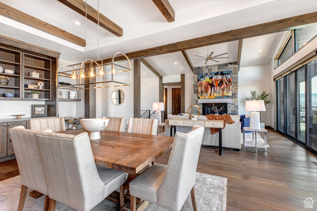 Dining space featuring lofted ceiling with beams, ceiling fan with notable chandelier, dark wood-type flooring, and a stone fireplace