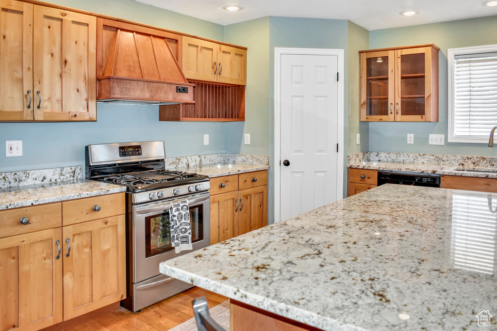 Kitchen with light stone countertops, premium range hood, stainless steel gas range oven, black dishwasher, and light hardwood / wood-style flooring