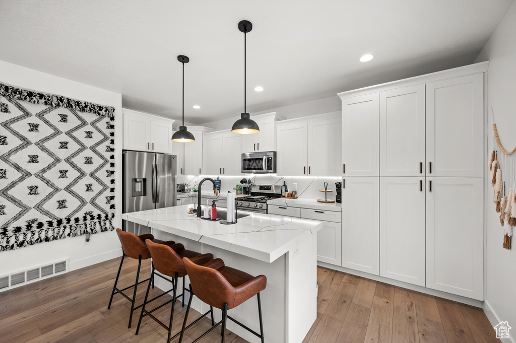 Kitchen featuring stainless steel appliances, white cabinetry, a center island with sink, light hardwood / wood-style flooring, and decorative light fixtures