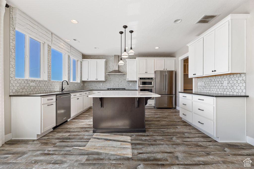 Kitchen with wall chimney range hood, appliances with stainless steel finishes, a kitchen breakfast bar, hanging light fixtures, and a center island