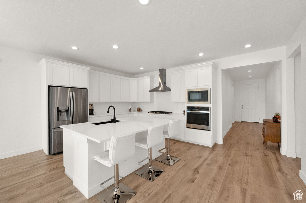 Kitchen featuring a kitchen island with sink, sink, light hardwood / wood-style floors, wall chimney exhaust hood, and appliances with stainless steel finishes