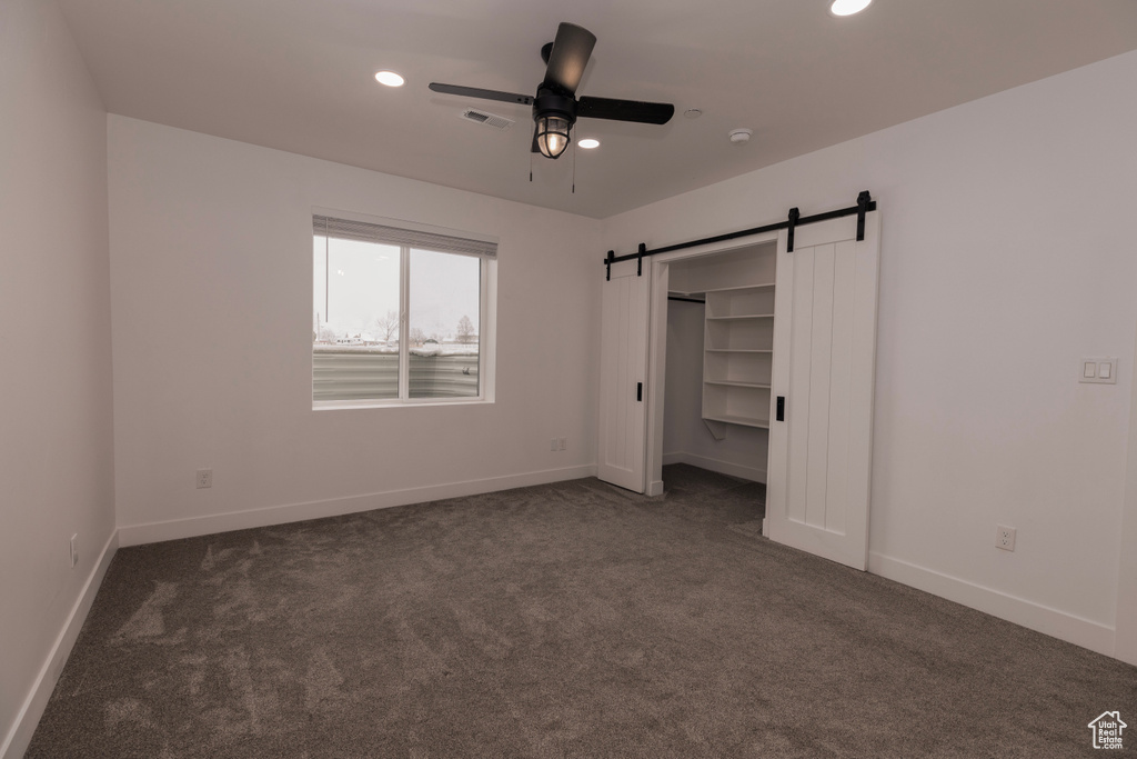 Unfurnished bedroom featuring dark colored carpet, a barn door, a closet, and ceiling fan
