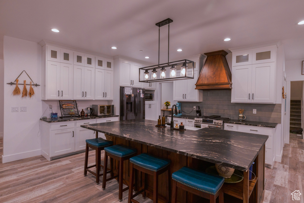 Kitchen with custom exhaust hood, a kitchen island with sink, appliances with stainless steel finishes, decorative light fixtures, and white cabinets