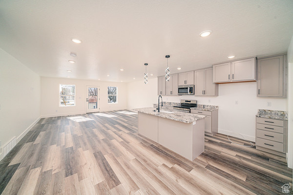 Kitchen with an island with sink, appliances with stainless steel finishes, light wood-type flooring, light stone counters, and hanging light fixtures