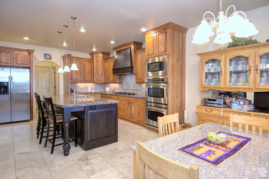 Kitchen with a notable chandelier, a kitchen island with sink, premium range hood, appliances with stainless steel finishes, and light stone counters