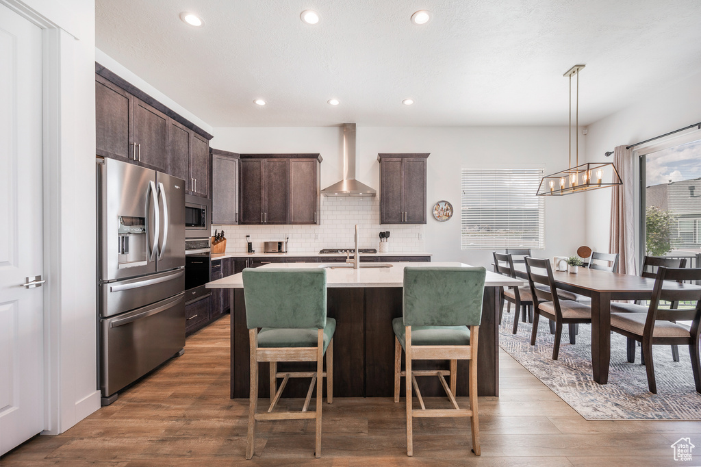 Kitchen featuring decorative light fixtures, wall chimney exhaust hood, backsplash, appliances with stainless steel finishes, and a center island with sink