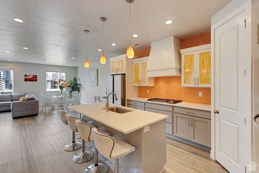 Kitchen with a kitchen island with sink, sink, custom exhaust hood, light hardwood / wood-style floors, and backsplash